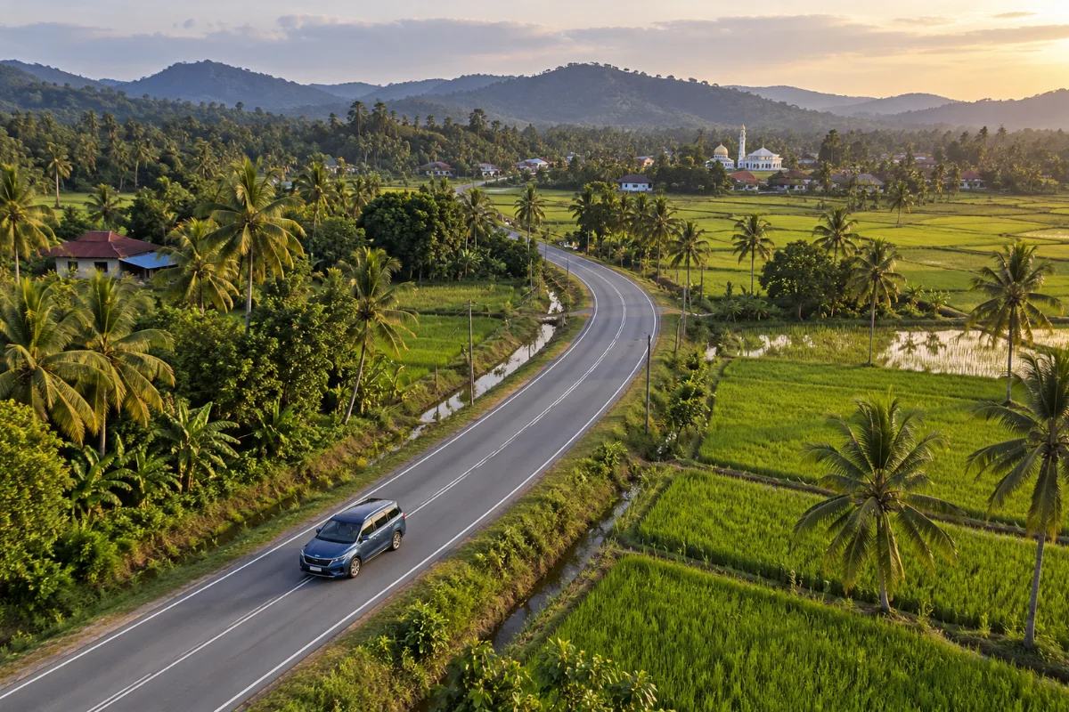 Blue Kia Carnival MPV seen from above driving on a quiet two-lane road through green Malaysian countryside at golden hour, a balik kampung car rental Malaysia trip