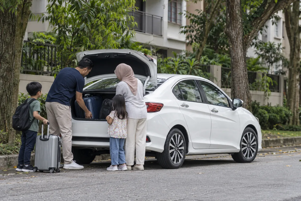 Malaysian family loading suitcases into the boot of a white Honda City Hybrid on a Kuala Lumpur residential street before a road trip